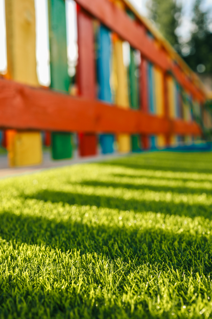 Artificial grass surface in a playground with colorful wooden fence, showing safe and durable turf option for children’s play areas.